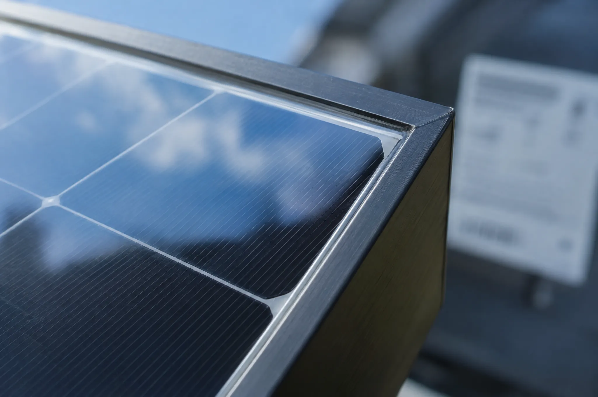 Imagem gerada por inteligência artificial: canto de um módulo solar monocristalino visto de perto, com moldura de alumínio escura e o vidro sobre a grade fina das células refletindo céu azul e nuvens. Ao fundo, a etiqueta traseira aparece desfocada e ilegível.